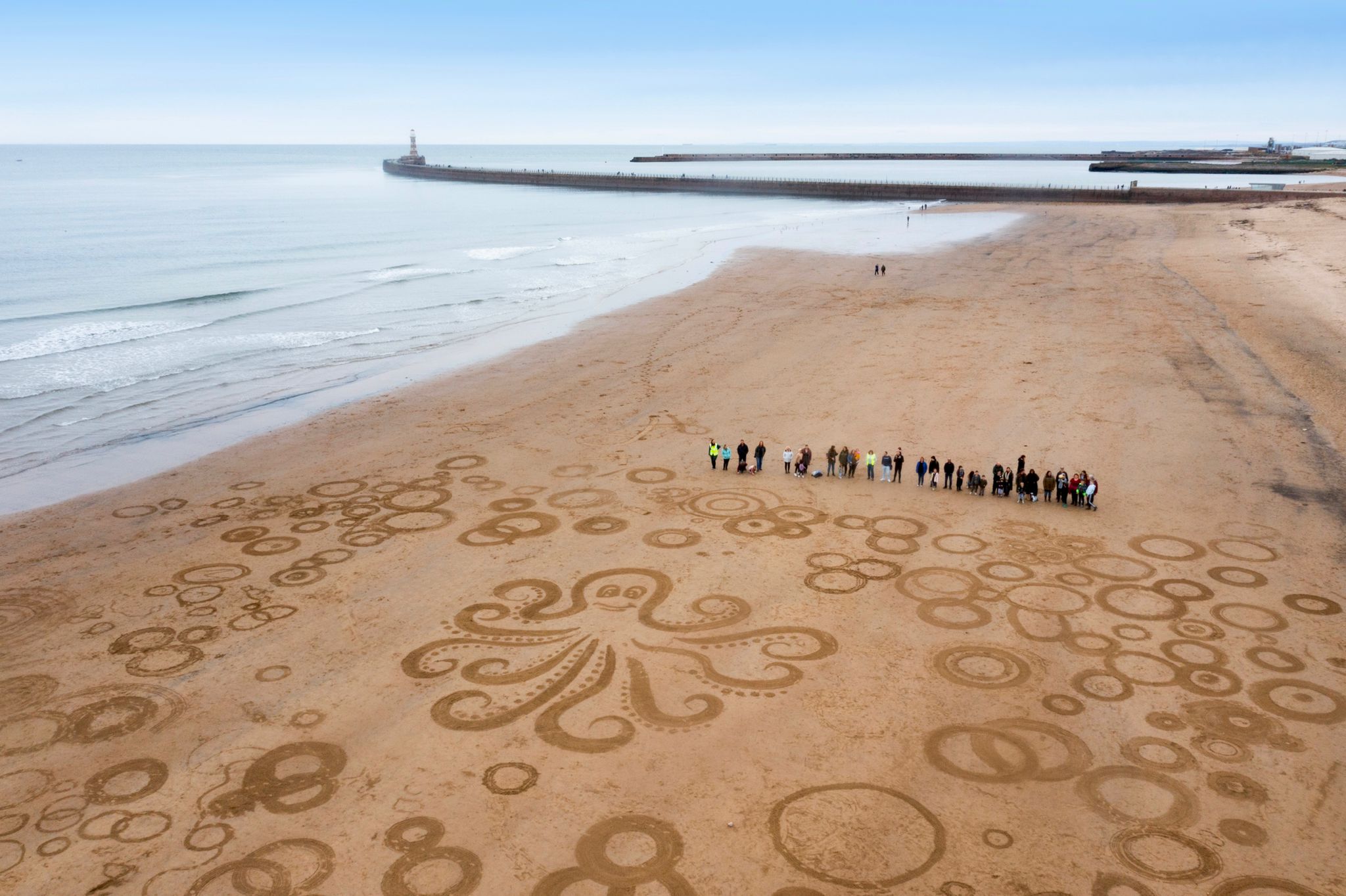 Families and staff from Adopt Coast to Coast gather around 100ft x 100fx octopus sand art with Roker lighthouse in the background